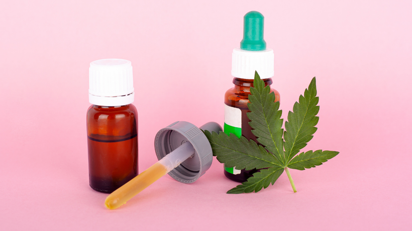 Cannabis leaf with amber dropper bottles and a syringe on a pink background