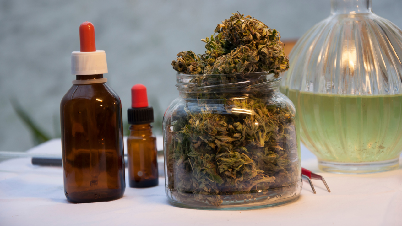Glass jar of cannabis buds with two amber dropper bottles and a glass vessel on a white surface