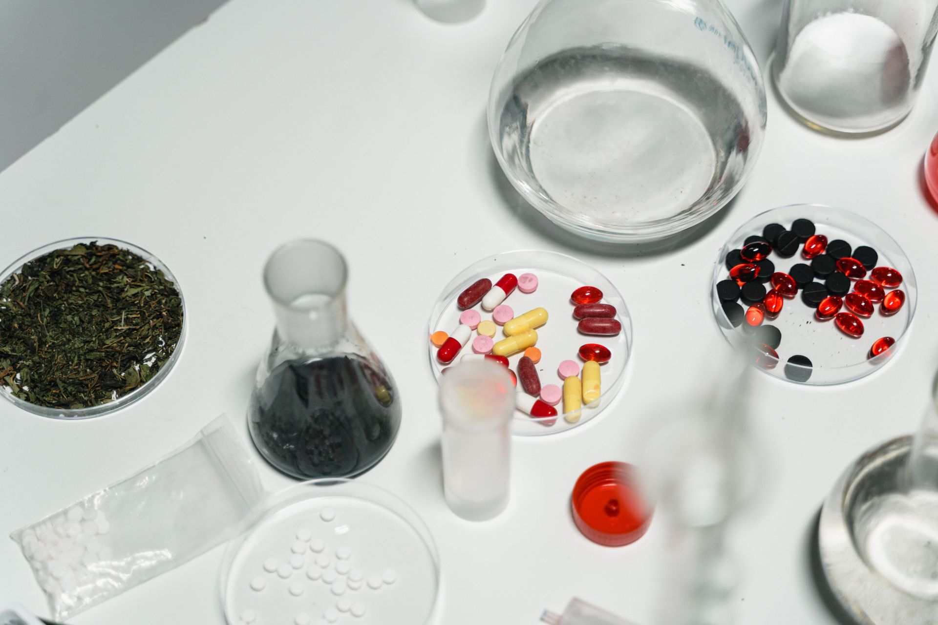 Assorted pills and capsules in petri dishes and small containers on a white lab table