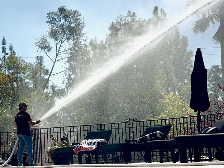 Person spraying a powerful water stream over a patio with chairs and an umbrella, trees in the background