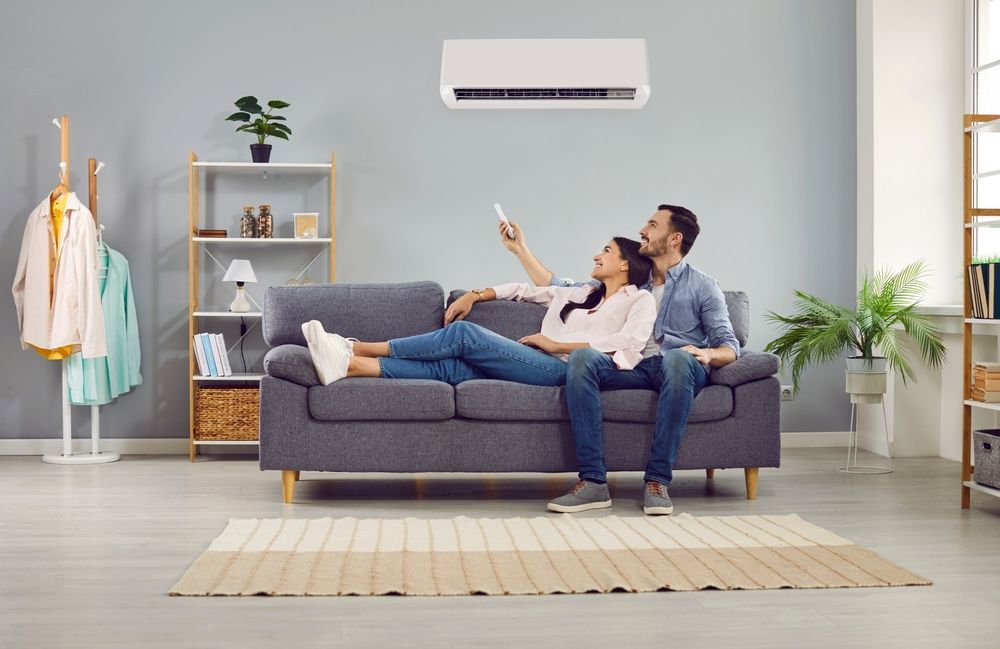 Couple relaxing on a gray sofa in a bright living room with a wall-mounted air conditioner.