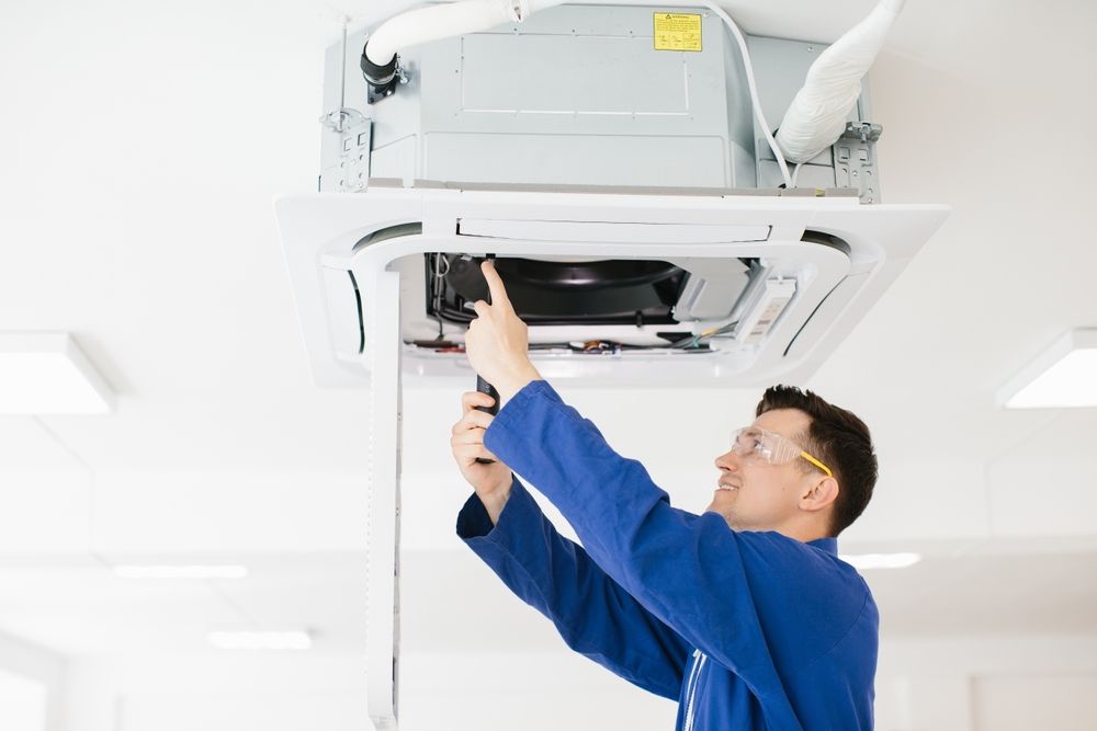 Technician in blue coveralls inspecting a ceiling-mounted air conditioner with a screwdriver