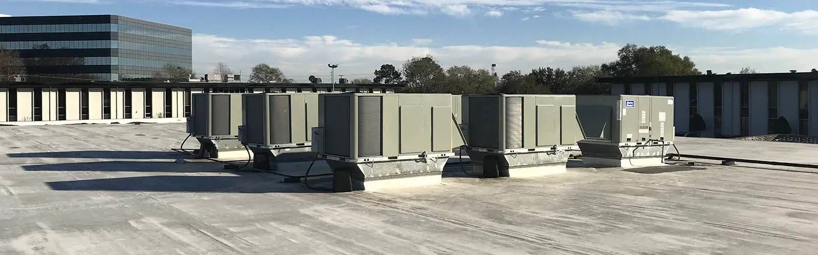 Rooftop with several beige HVAC units and ducts beside a glass office building under a cloudy sky