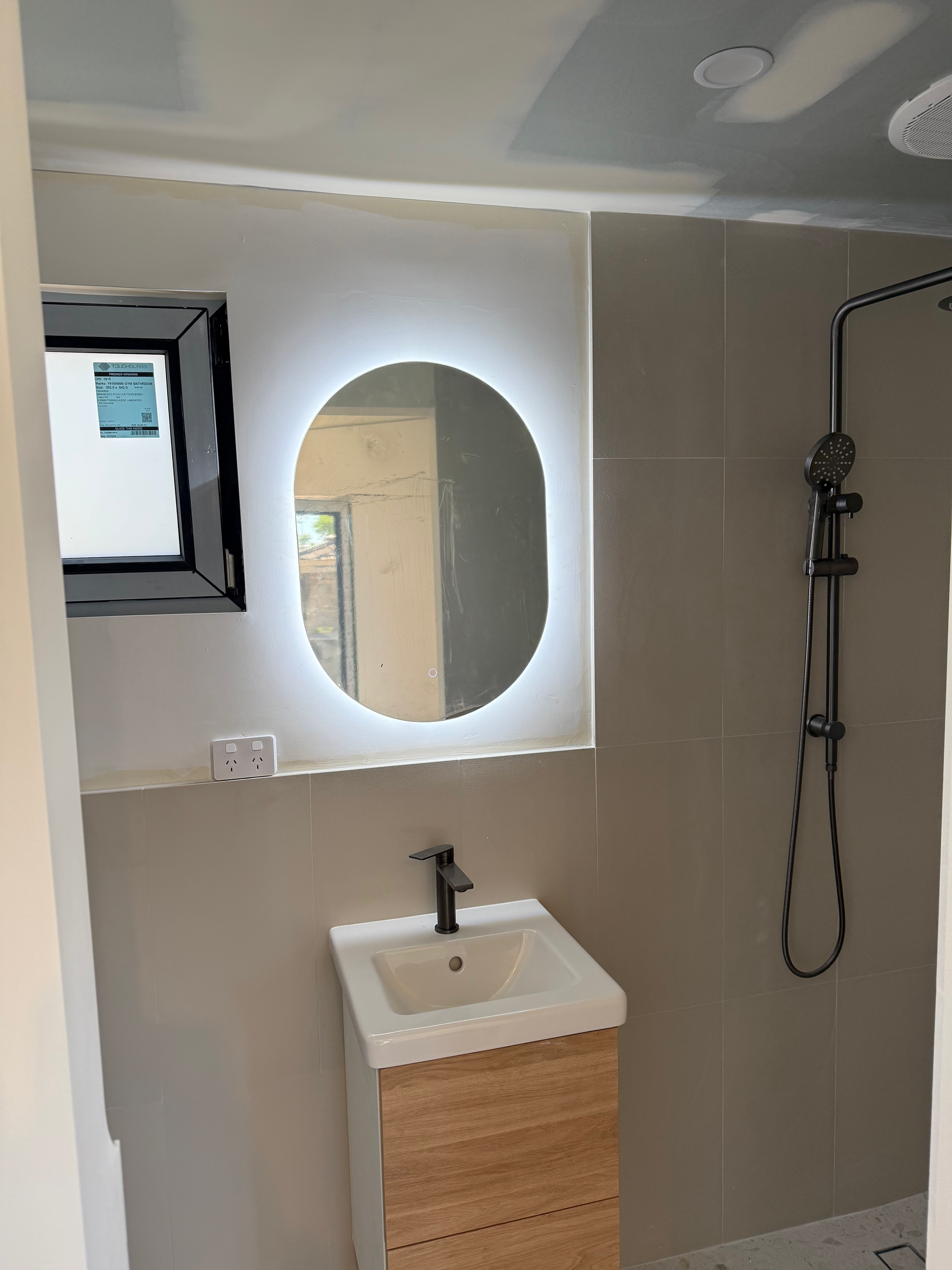 Modern bathroom with lit oval mirror, wood vanity, window, and black shower fixture.