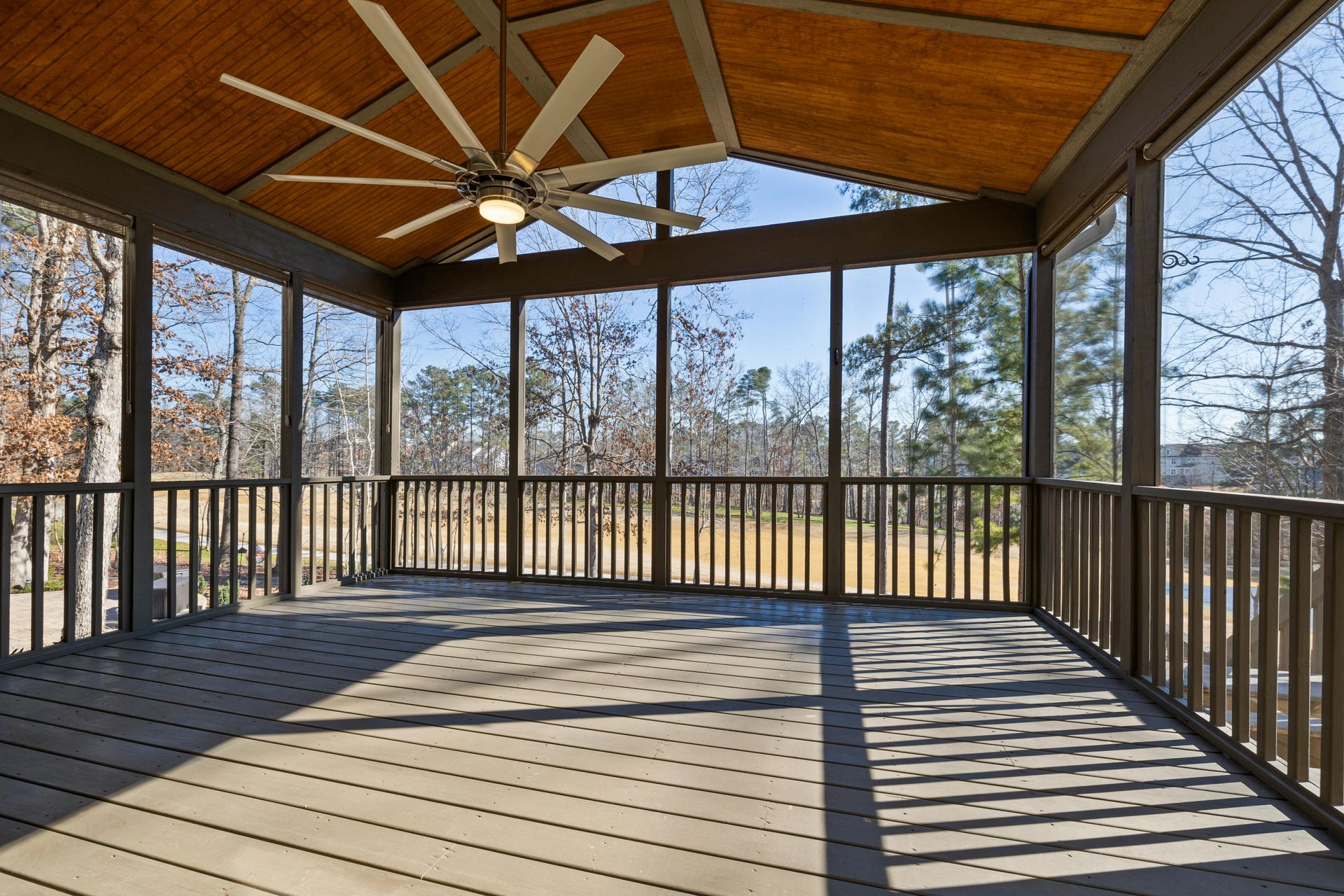 Covered wooden deck with railings, vaulted wood ceiling, and ceiling fan, overlooking bare trees.