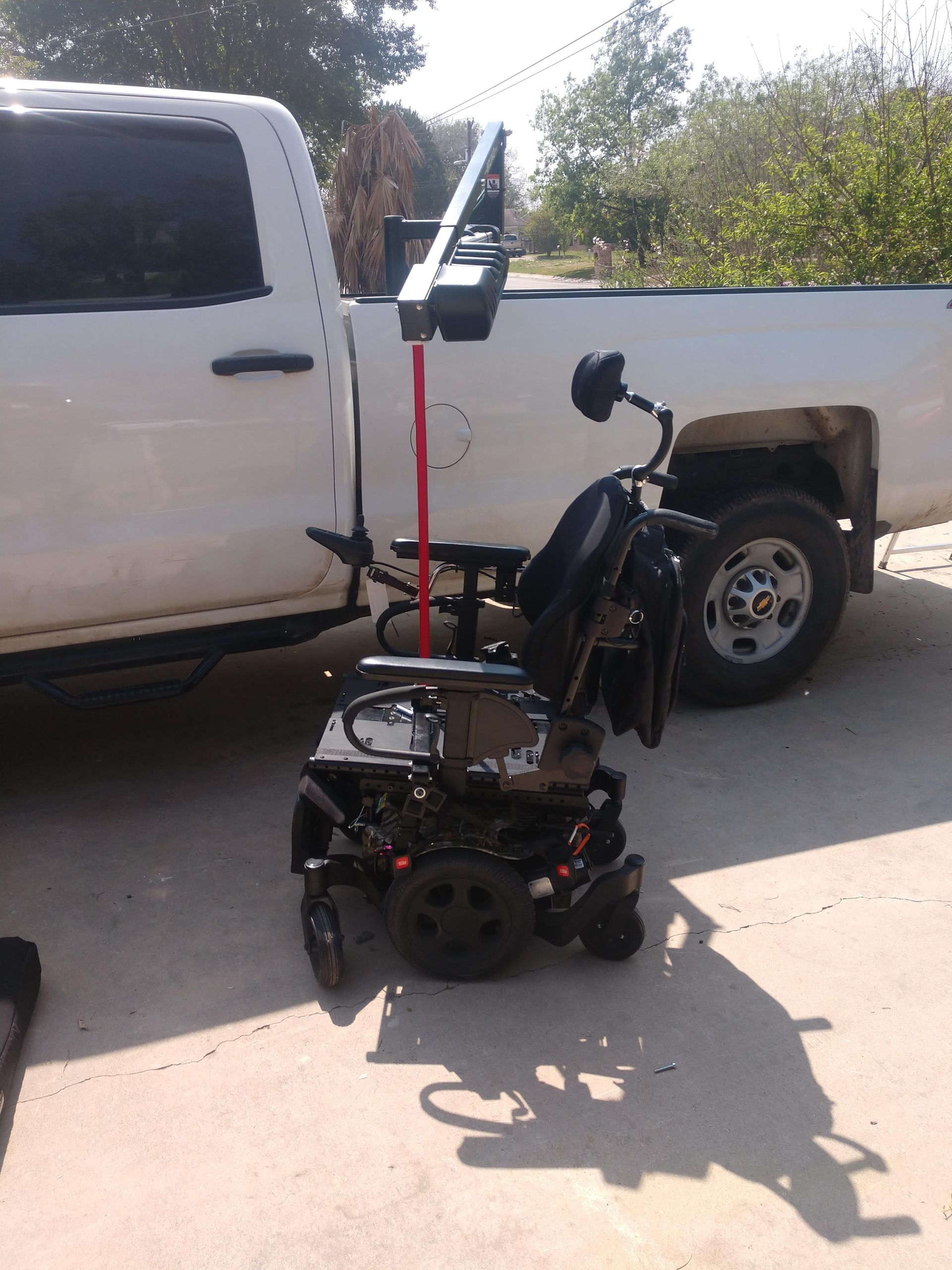 Black motorized wheelchair next to a white pickup truck, with a red pole and arm extending upwards.