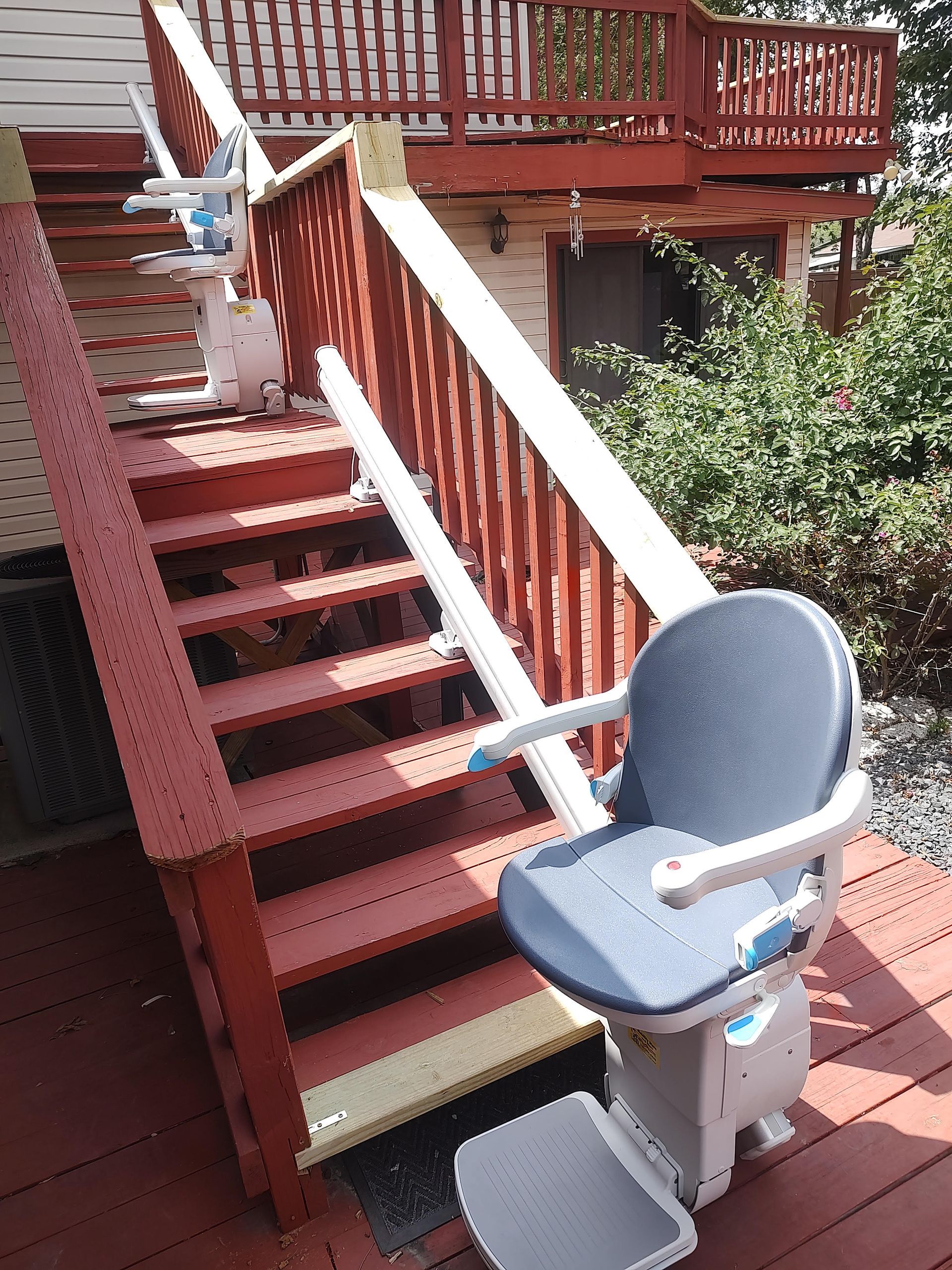 Stair lift on outdoor deck with wooden stairs and red railings. Blue chair, cream support, green shrubs.