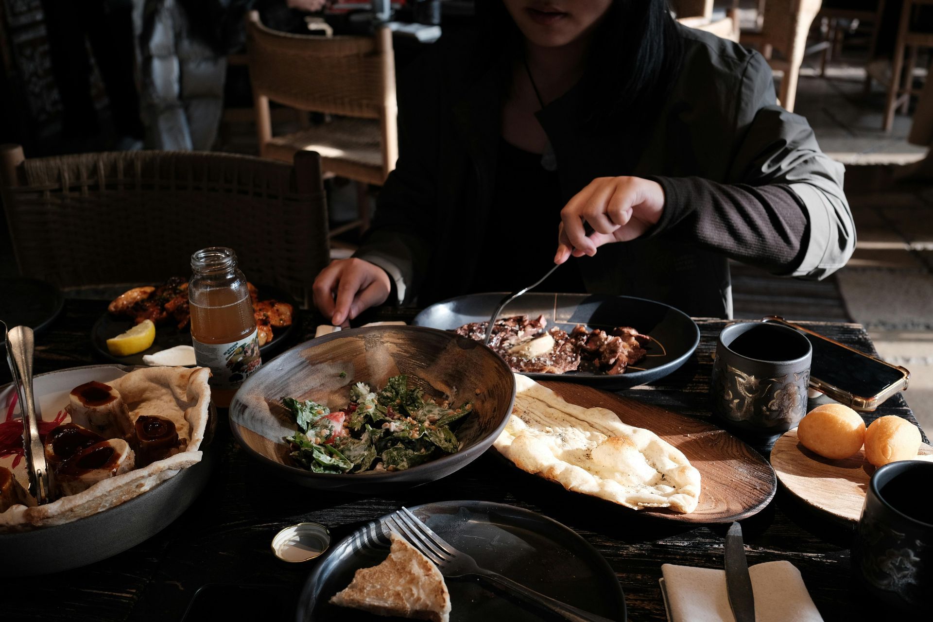Person eating a meal at a table with various dishes: salad, bread, beans, and drinks in a restaurant setting.
