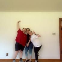 Three people posing together in a bare room, smiling and leaning close to the wall.