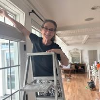 Woman standing on a step ladder indoors, reaching up near a window in a bright living room.