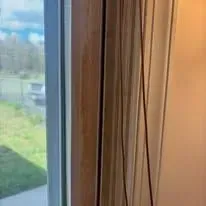 Window with beige curtains beside a view of green grass and a driveway outside.