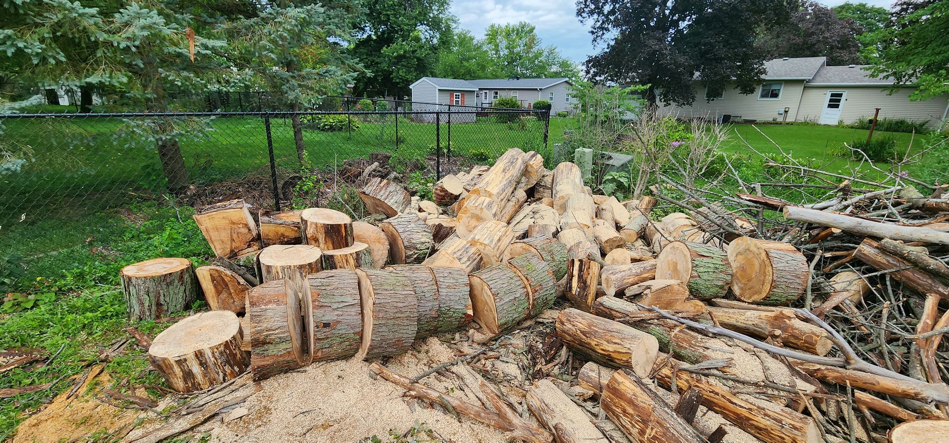 A pile of logs and stump in a yard with a house in the background.
