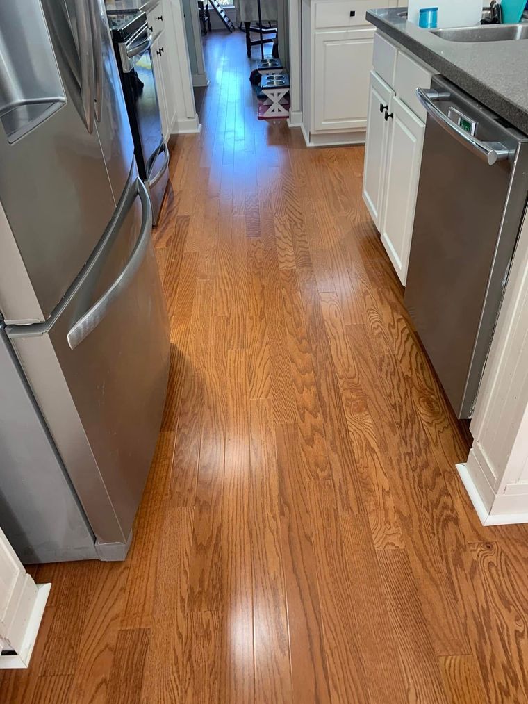 Narrow kitchen hallway with wood floors, stainless steel fridge, and white cabinets leading to a bright back room