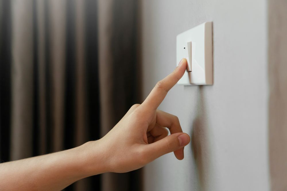 Hand pressing a light switch on a white wall. Grey curtain in the background — Holders Electrical In Inverell, NSW