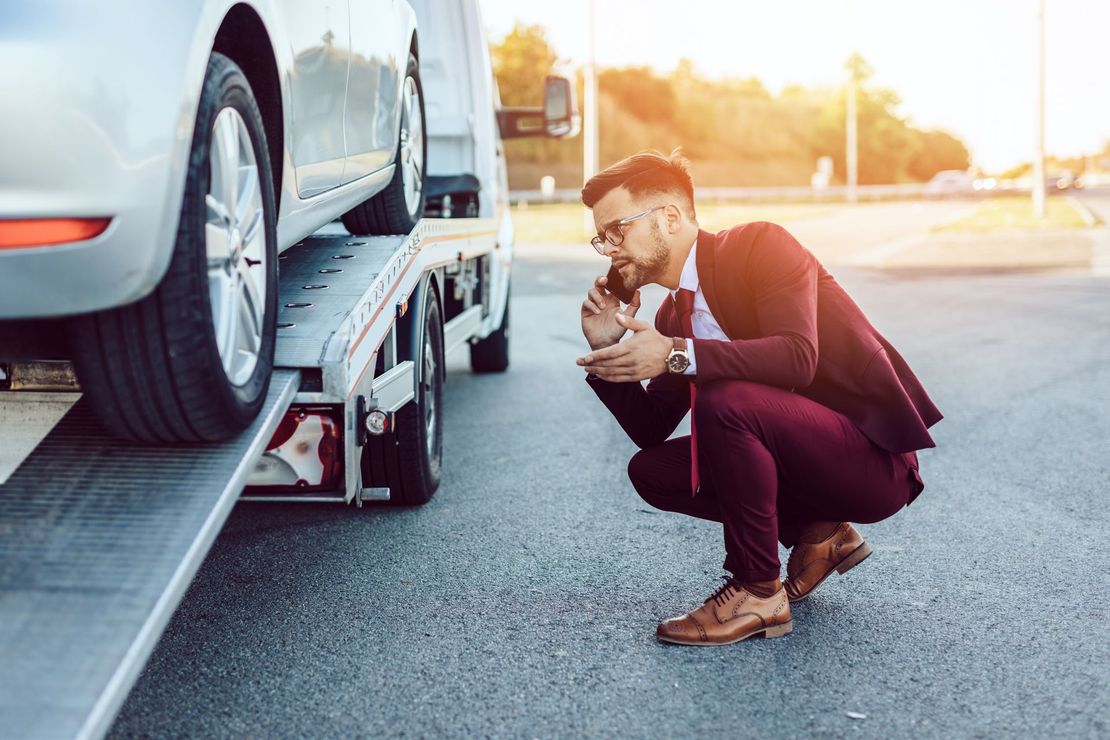 Man crouching beside a car being loaded onto a tow truck, inspecting the front wheel on a sunlit street