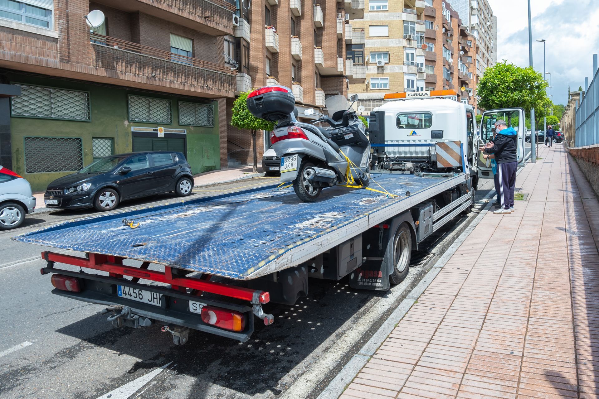 Tow truck loading a scooter on a flatbed along a city sidewalk.