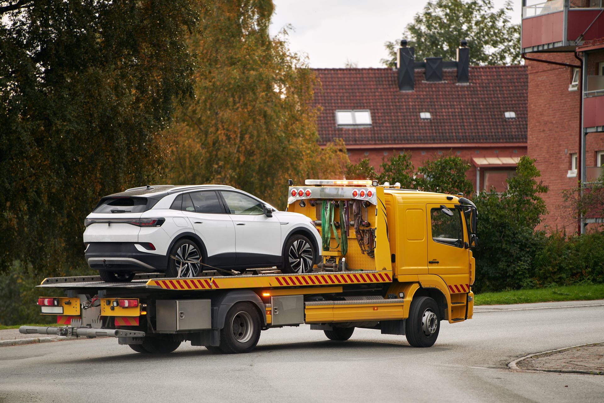 Yellow tow truck carrying a white SUV on a street beside houses and trees