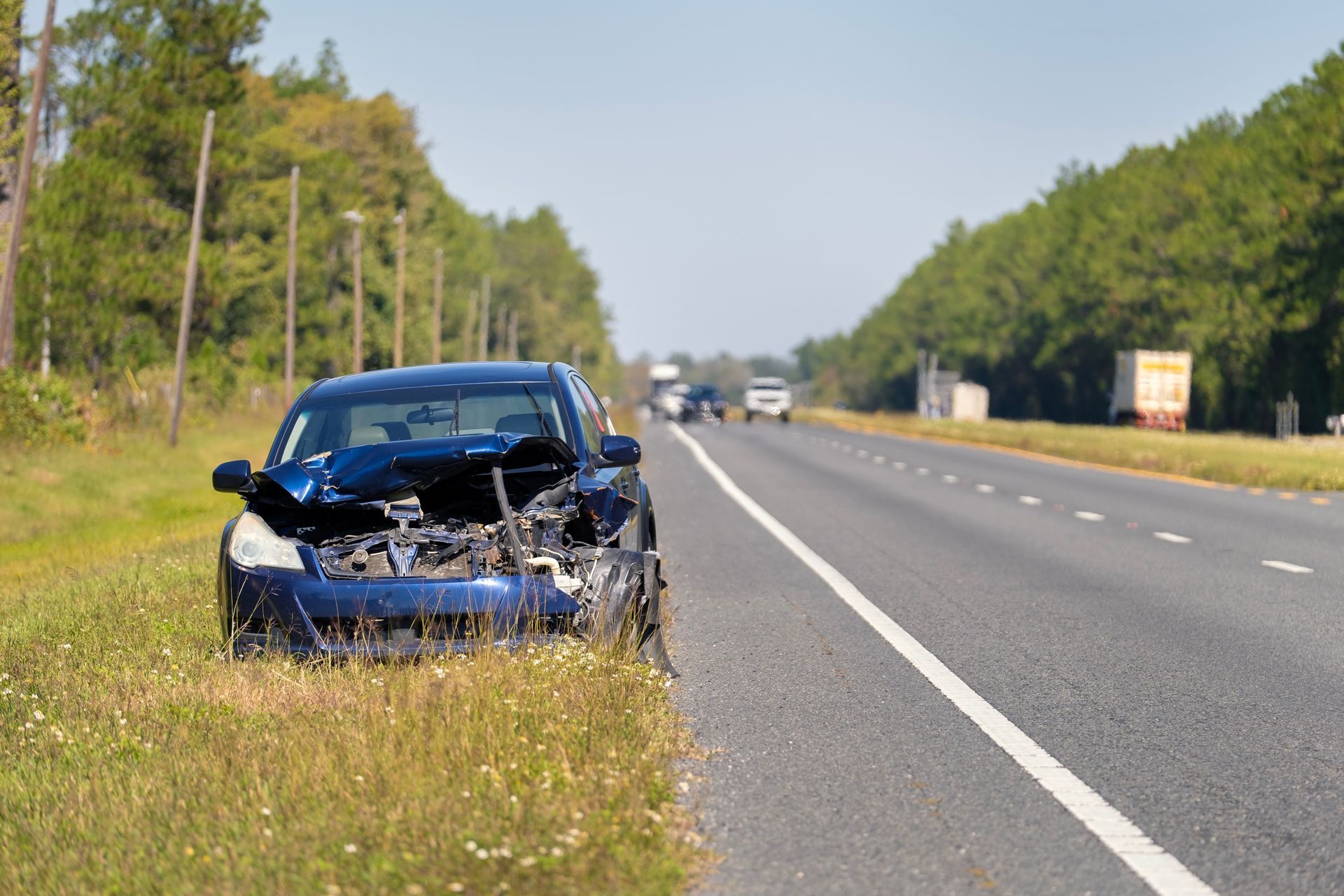 Damaged black car on the roadside beside a highway, with front-end crash damage and no people visible