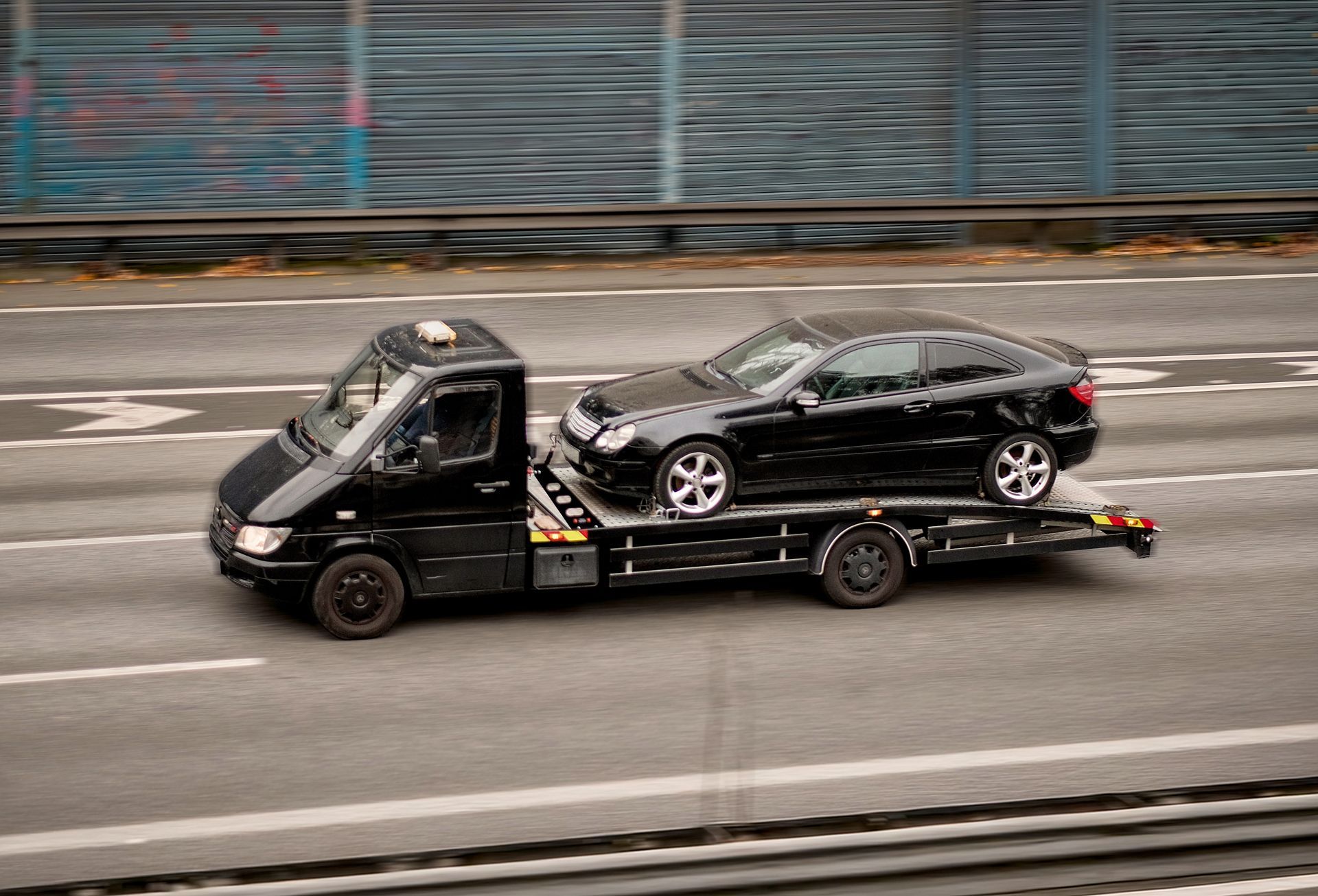 Black car on a flatbed tow truck driving on a city street.