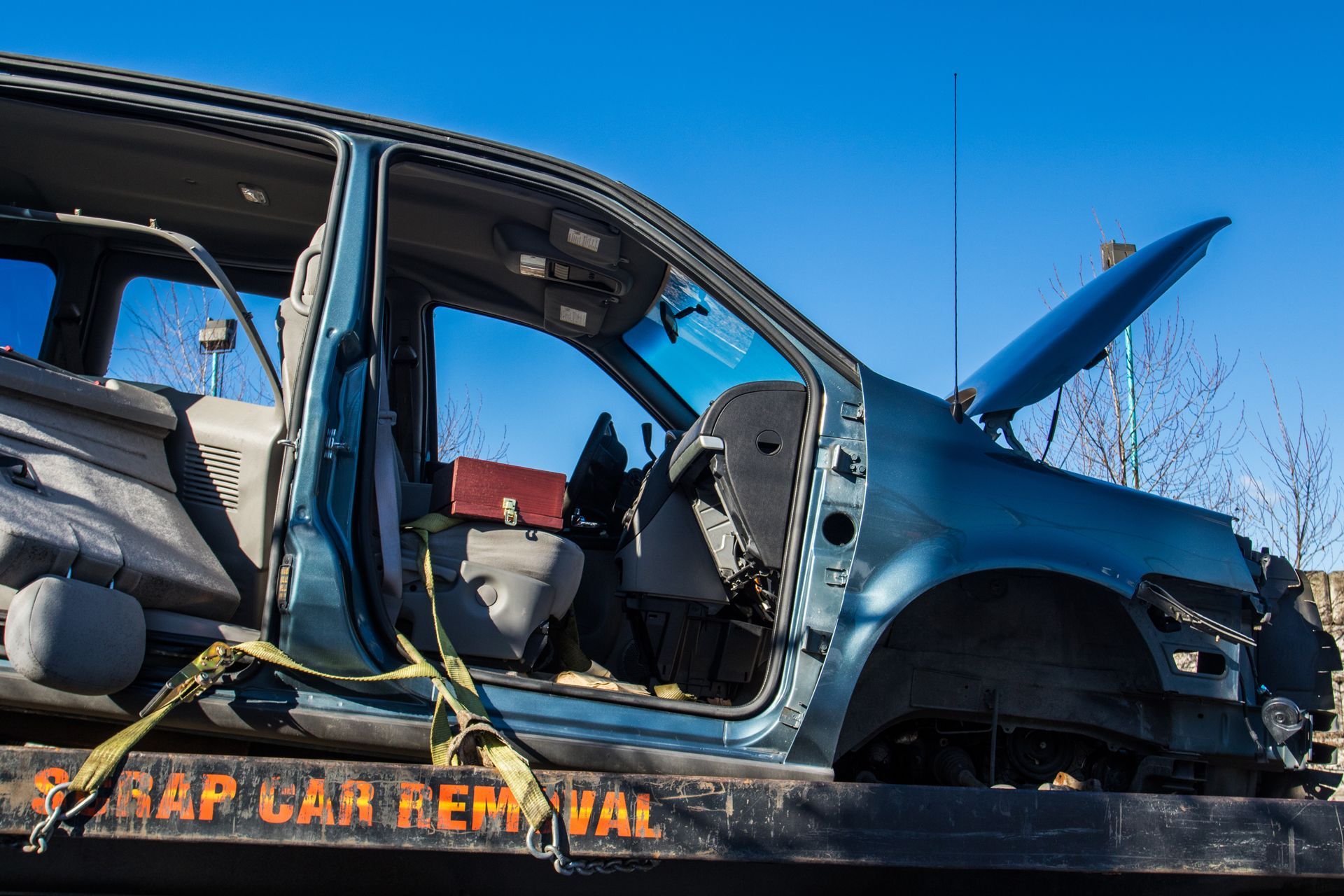 Damaged blue car on a tow truck, with the hood open and missing doors, at a scrapyard