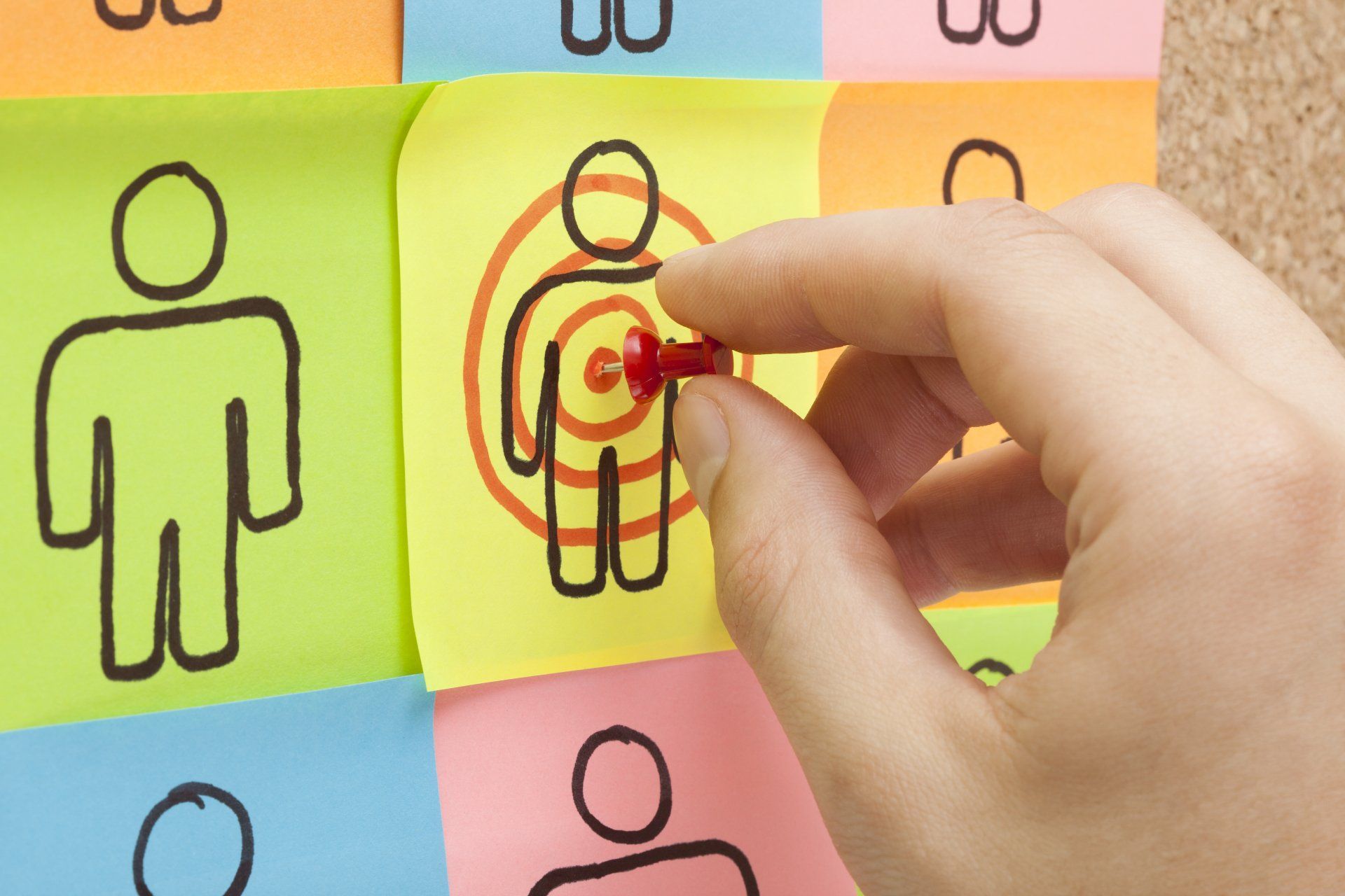 Hand placing a red pushpin on a sticky note with a person symbol inside a target; other notes with people.