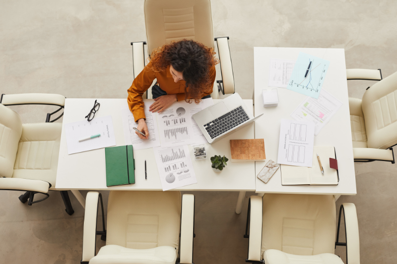 A person in an orange top works at a desk with a laptop, documents, and office supplies, viewed from directly above.