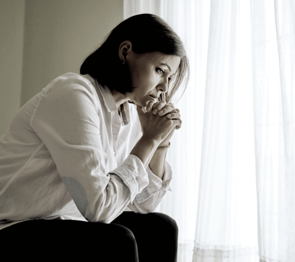 A woman is sitting in front of a window with her hands folded in prayer.