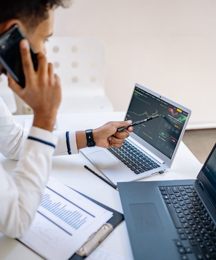Trader on phone points at laptop with stock chart on desk in bright office