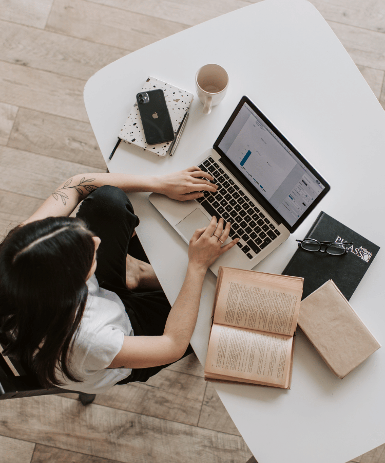 Person working on a laptop at a white table with books, notebook, and coffee cup