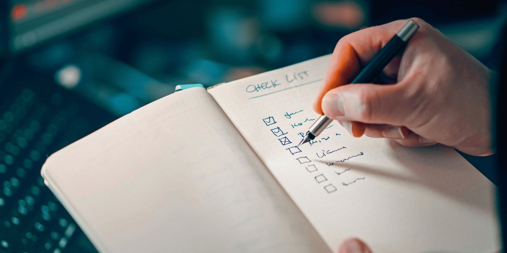 Hand writing a checklist in a notebook beside a laptop on a desk
