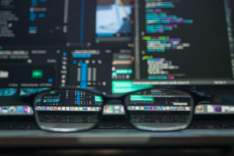Eyeglasses on a laptop reflect coding on the screen; blurred background of code on a monitor.