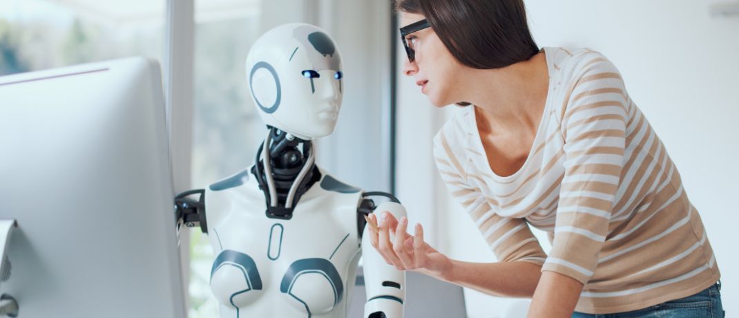 A woman in glasses talks to a white robot near a computer in a bright room.