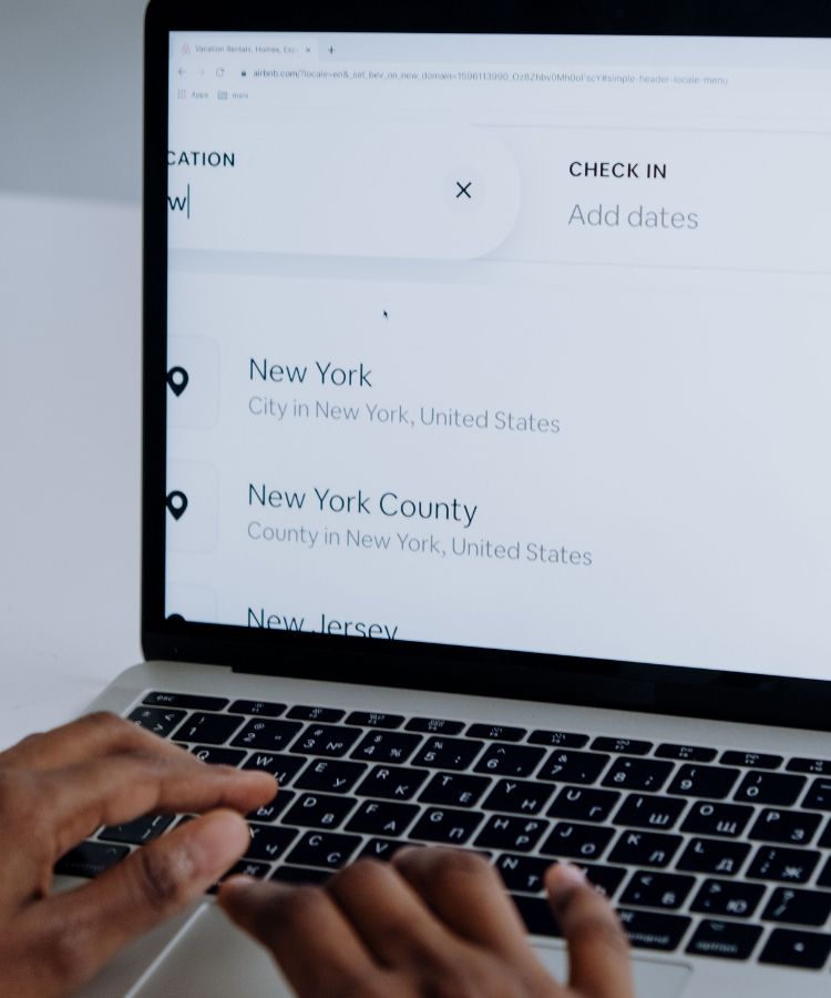 Hands typing on a laptop showing a travel search page with New York locations and check-in dates.