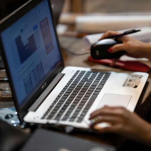Person using a laptop, data charts on screen. Mouse and pen in hand, working at a table.
