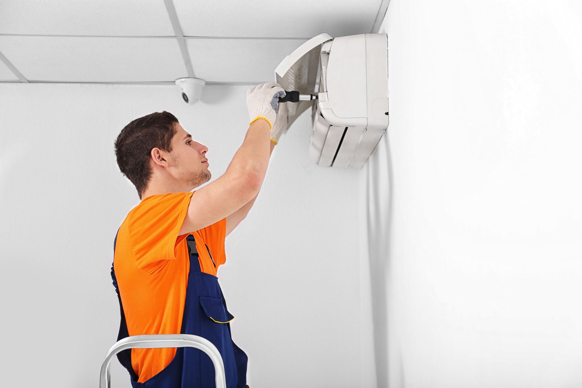 Technician in orange shirt and blue overalls repairing a wall-mounted air conditioner indoors