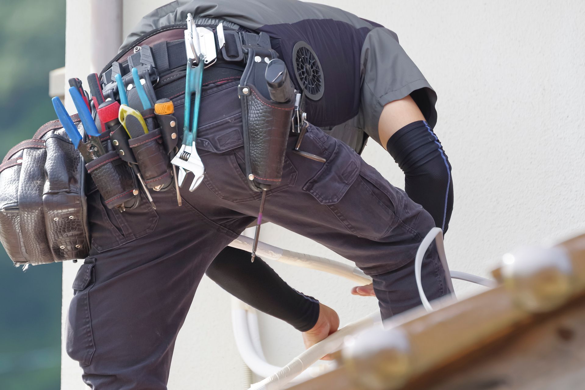 Roofer crouching on a roof, wearing a tool belt and using a white cable or hose.