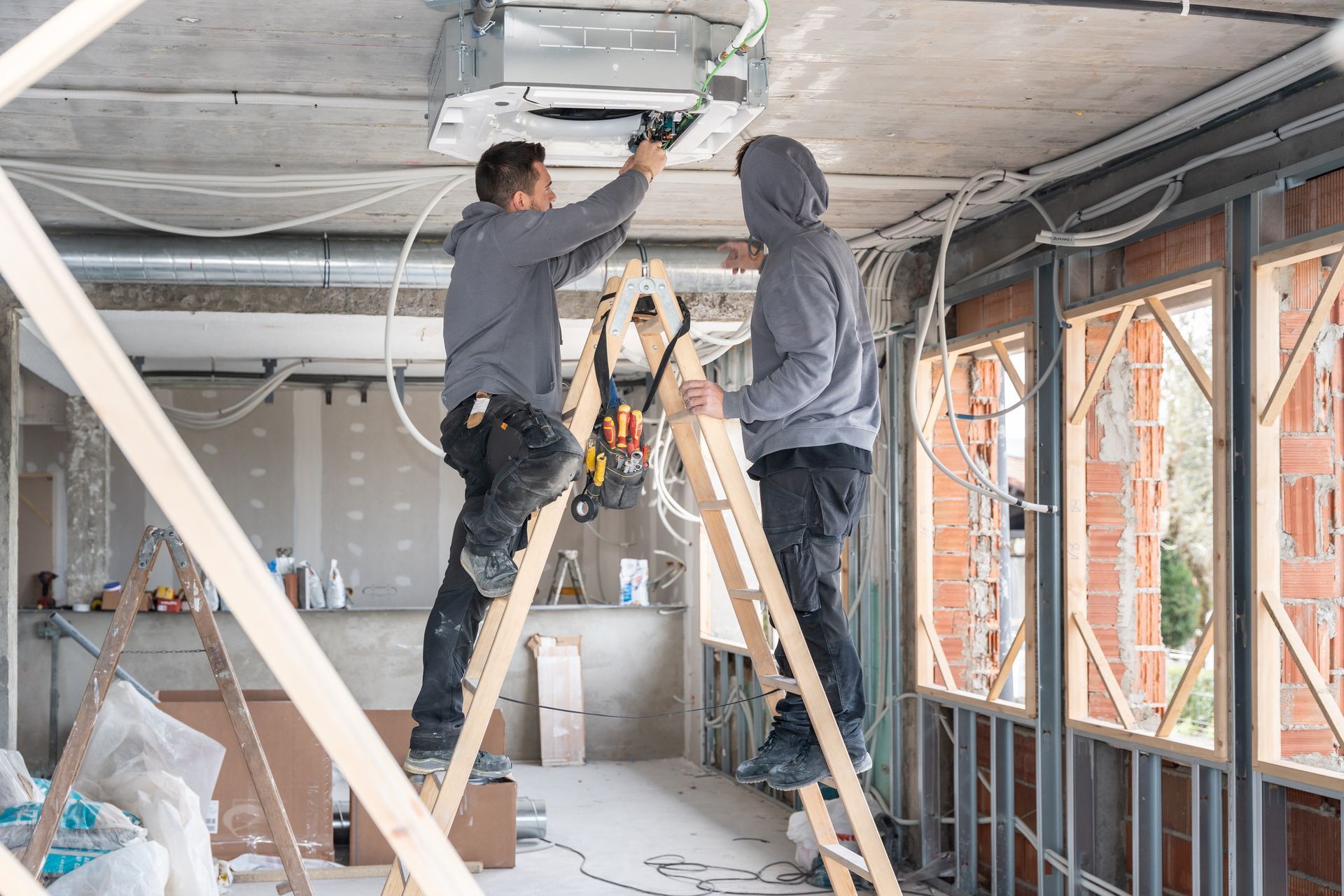 Two workers on ladders installing a ceiling fixture in a room under construction.