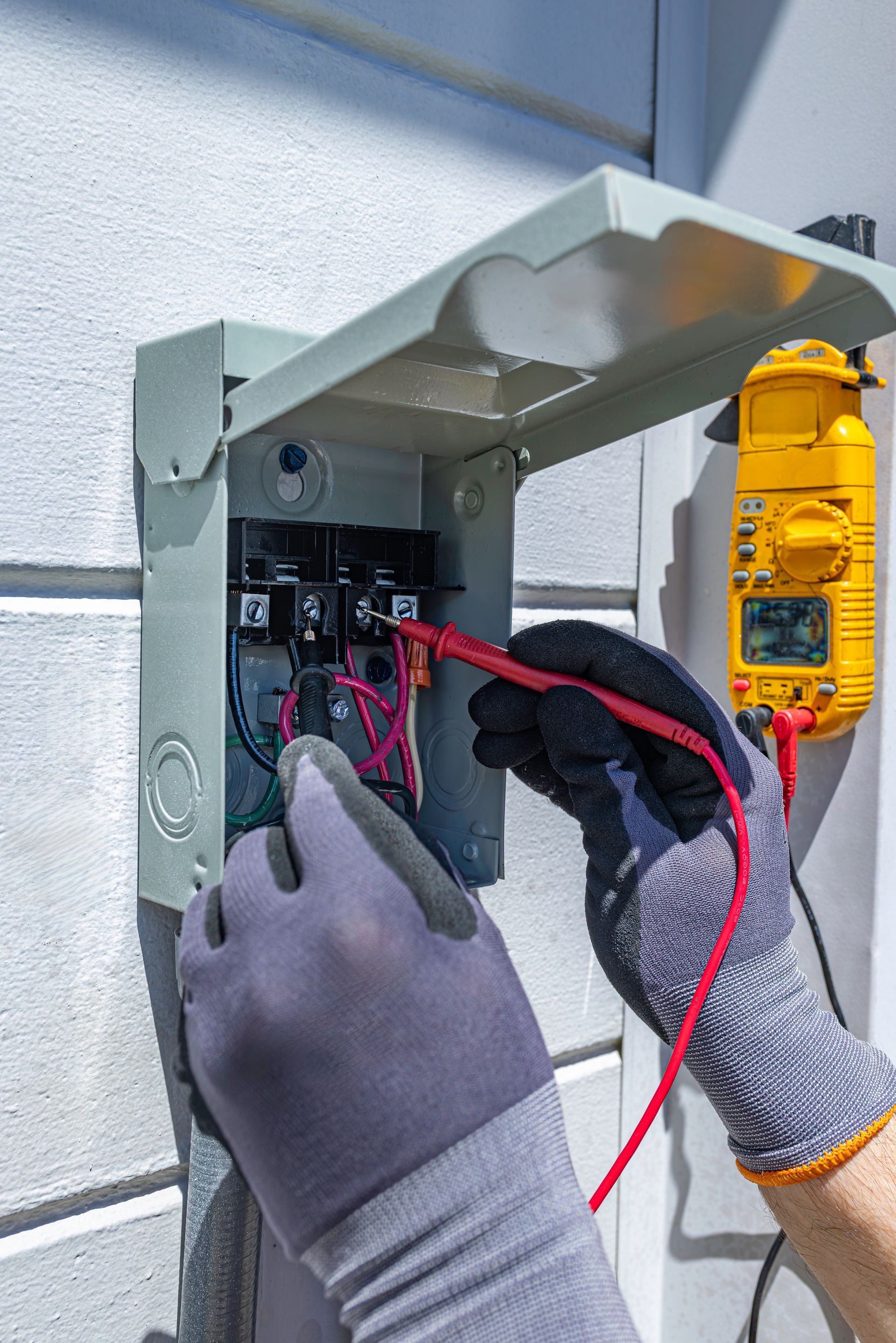 Gloved hands wiring an outdoor electrical panel with a multimeter on the right side.