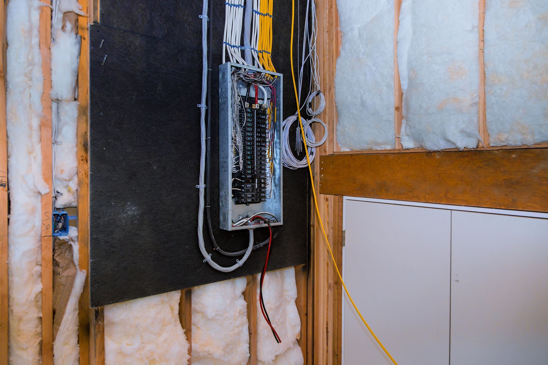 Electrical panel with exposed wiring mounted on an unfinished wall beside a white cabinet