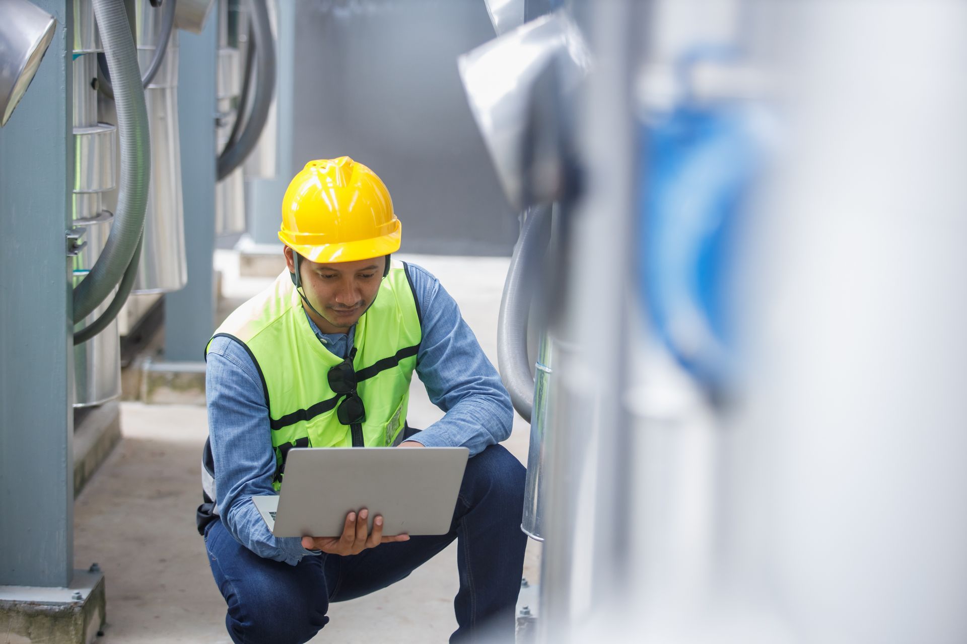 Worker in yellow hard hat kneeling and using a laptop in an industrial facility