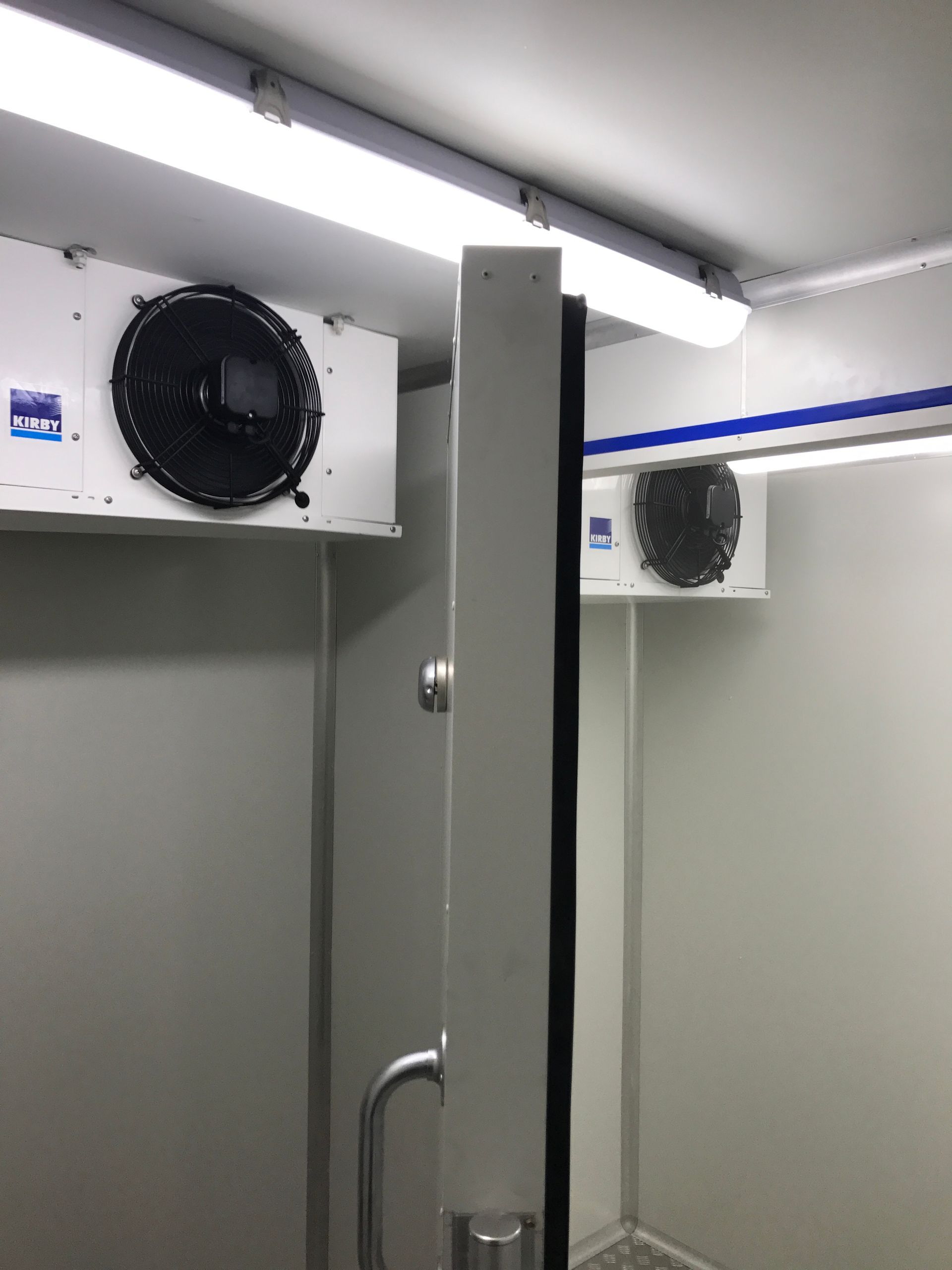Public restroom shower stalls with gray partitions and wall-mounted fans under fluorescent lights