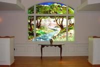 Bright room with an arched window framing a tropical stream and trees, above a small table on wood floor