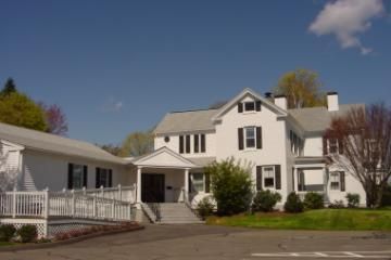 White house with black shutters and a covered porch beside a parking lot under a blue sky