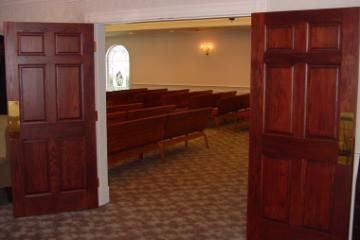 Open church doors leading to a pew-lined sanctuary with patterned carpet and warm lighting