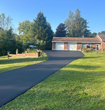 Long black asphalt driveway leading to a brick house with two garage doors. Lush green lawn surrounds.