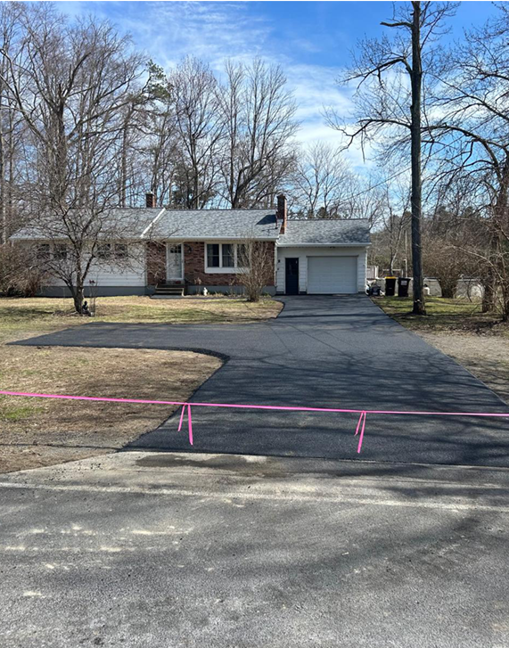 Asphalt driveway leading to a one-story house with a brick facade and attached garage; pink tape marks the edge of the driveway.