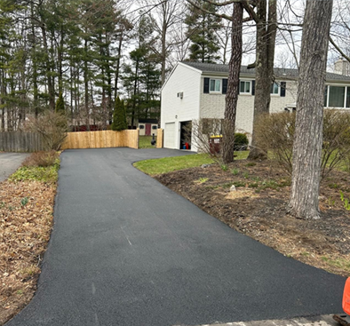 Black asphalt driveway leads to a two-story white house with a garage; trees and a wooden fence flank the drive.
