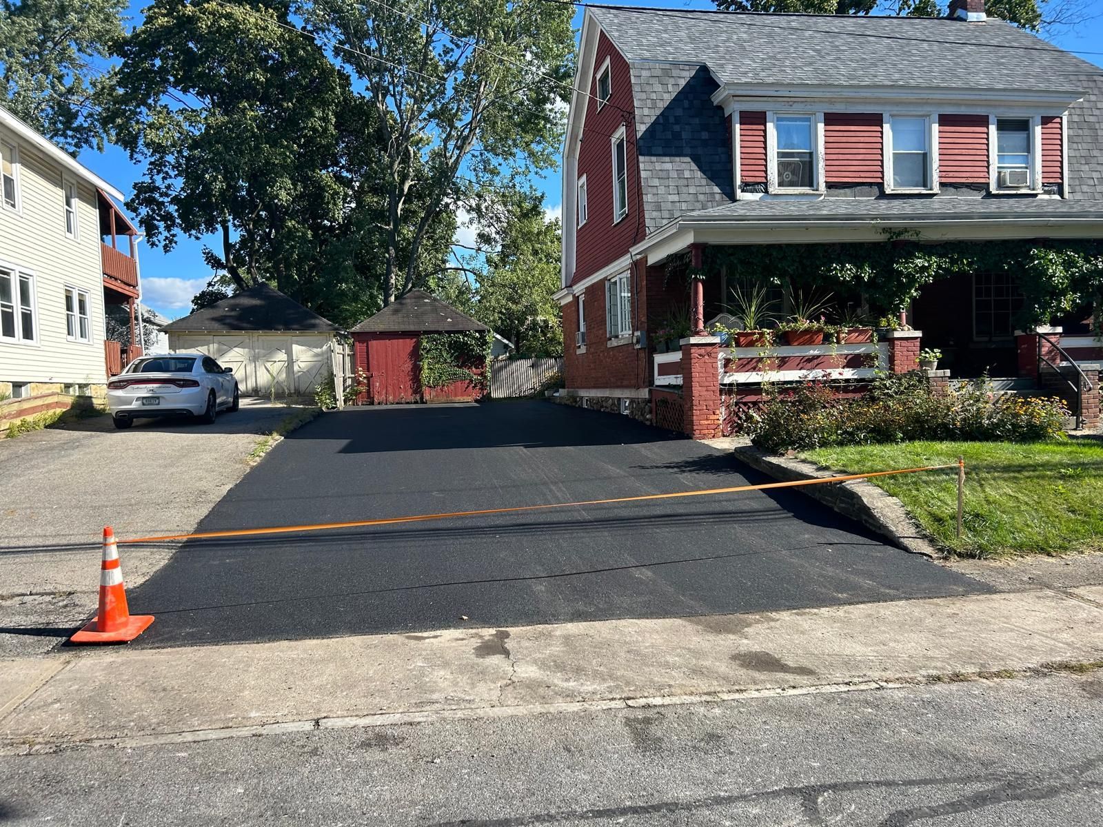 Freshly paved black driveway in front of a red brick house with porch, orange cone at edge.