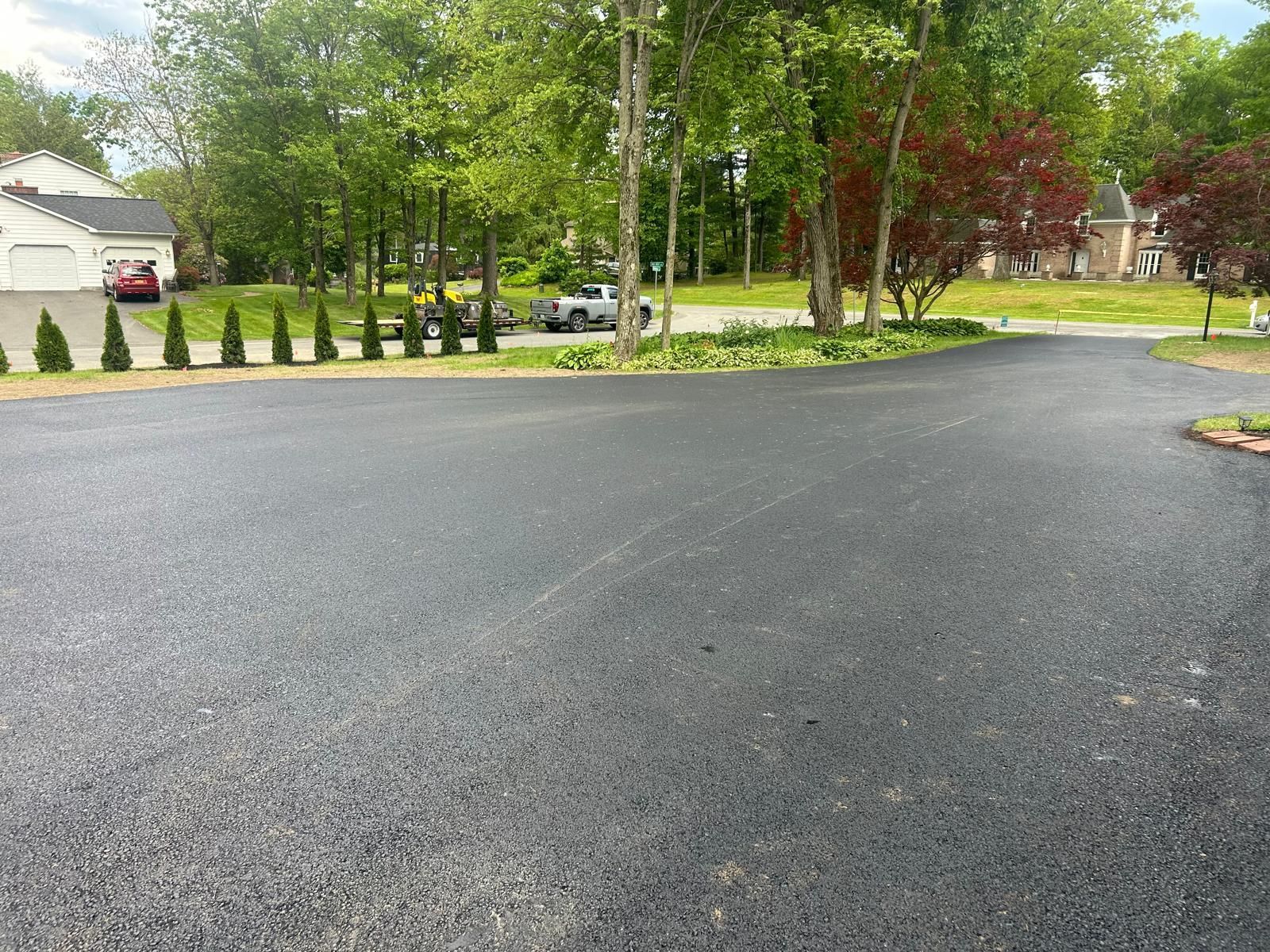 Newly paved asphalt driveway with line of trees and houses in the background.