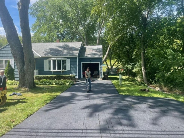 Black asphalt driveway leads to a blue house with garage; person stands in the driveway. Green trees surround.